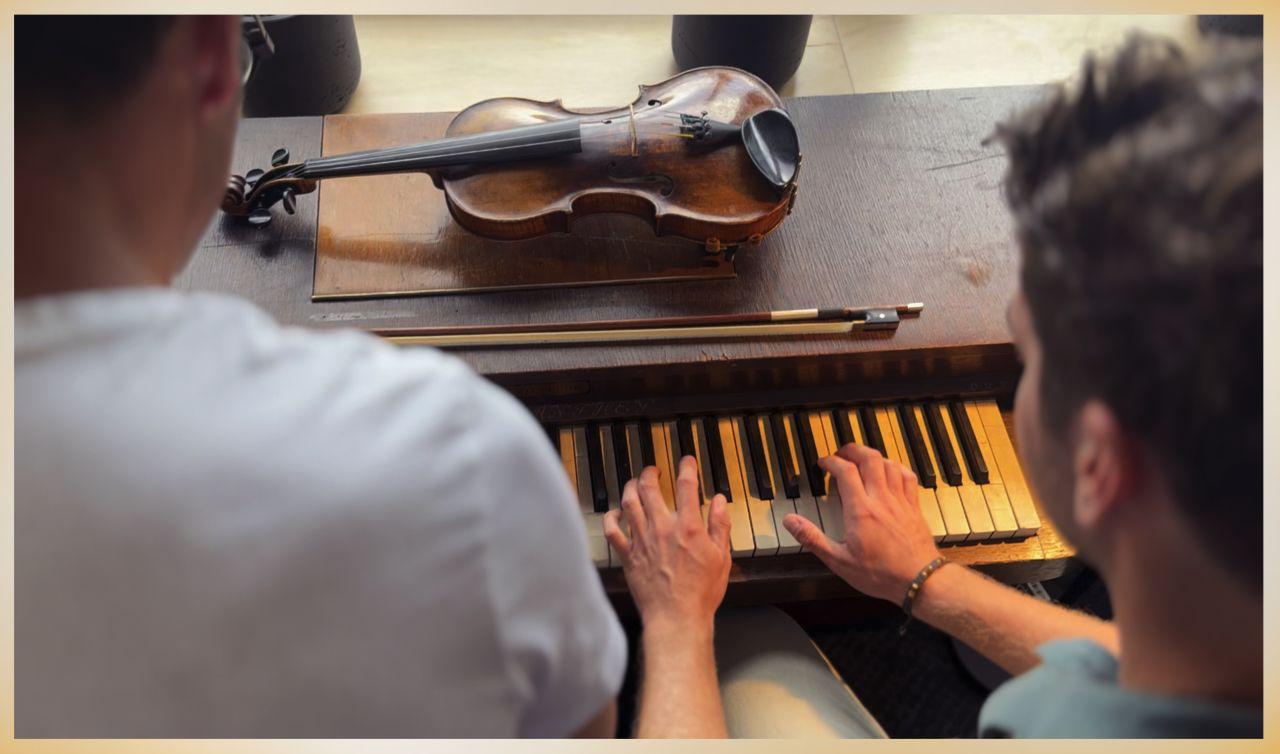 Two people sharing the keys of the upright piano, four hands on the keyboard, a violin and bow resting on the wood above them