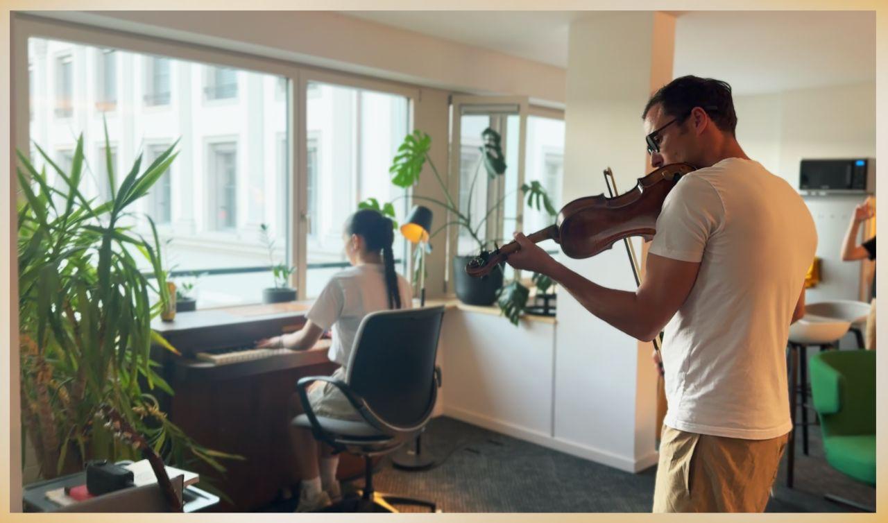 A violinist playing beside the windows at Ship26 while a woman with a long braid accompanies him on the upright piano, plants and desks around them