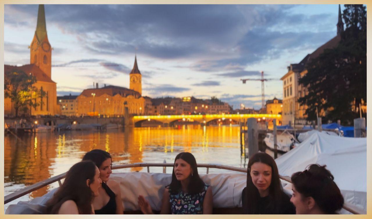 Five people talking in the stern of the boat as night falls, the Fraumünster spire, St. Peter's clock tower and a lit bridge reflected in the Limmat behind them