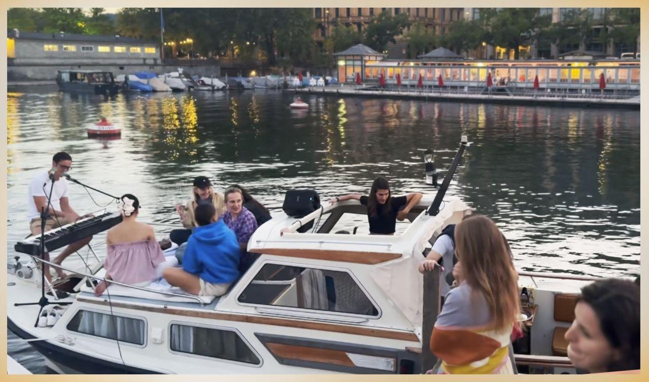 A musician playing an electric keyboard into a mic on the deck of the community boat at dusk, the crew sitting around him and the lit quay across the water