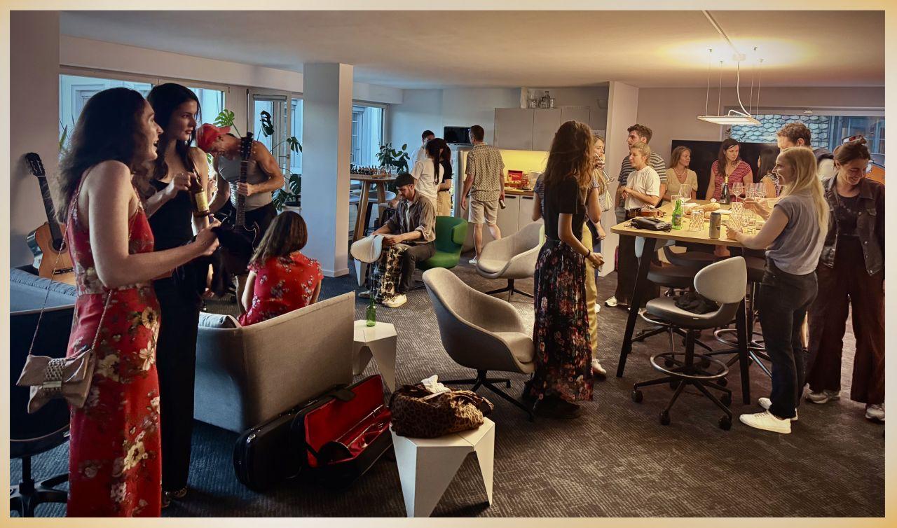 The room at Schifflände 26 full of people with drinks, a saxophone player and a guitarist mid-song, acoustic guitars on stands and an open violin case on the floor
