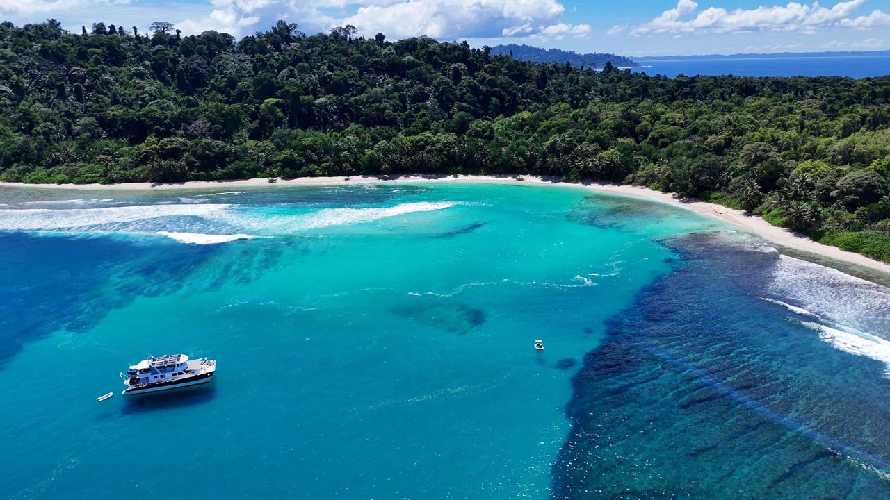 Drone shot of the surf charter anchored off a blue coast, an island rising behind it